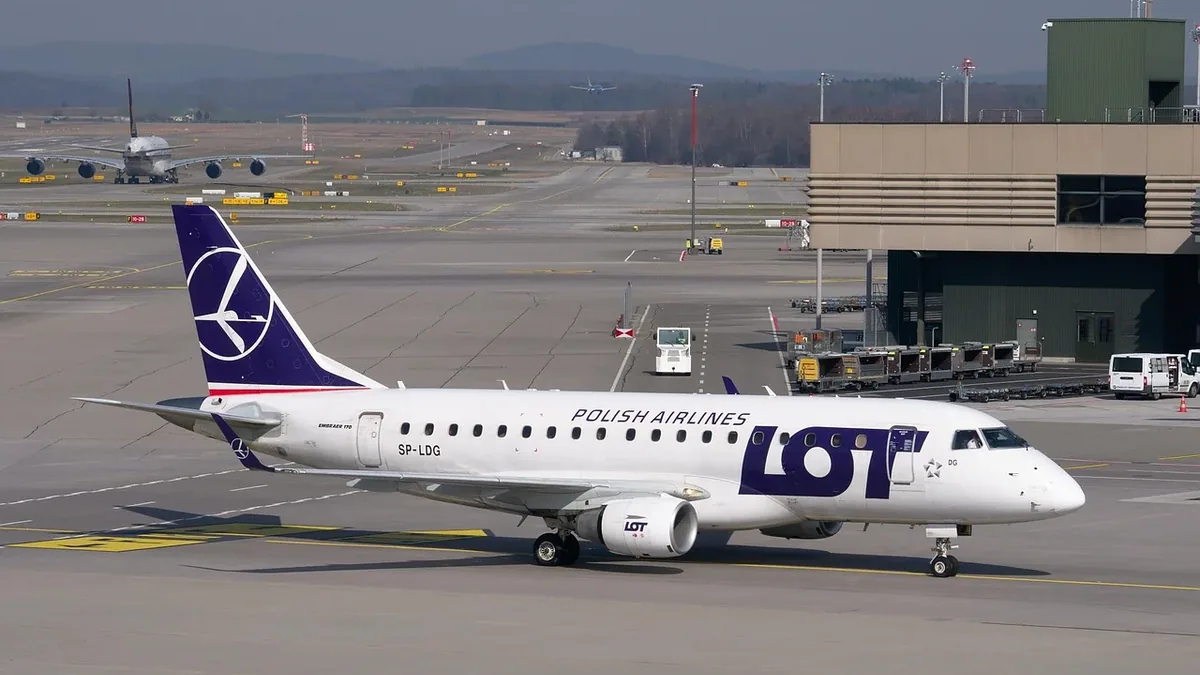 A white and purple Polish Airlines plane on a runway, with a building and other planes in the background.